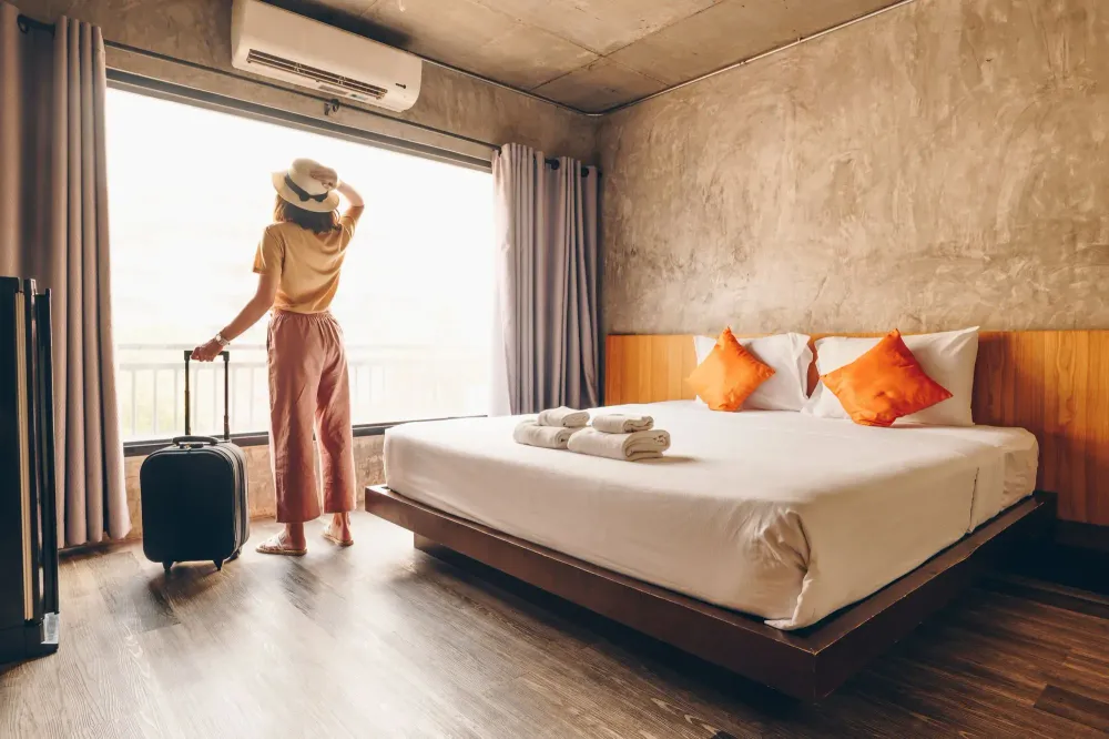 Woman standing in a hotel room and looking out the window, holding the handle of her luggage